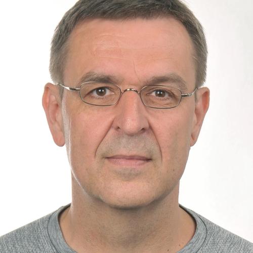  A man with short graying hair and glasses, wearing a light gray long-sleeved shirt, looks directly at the camera in a headshot against a plain white background.